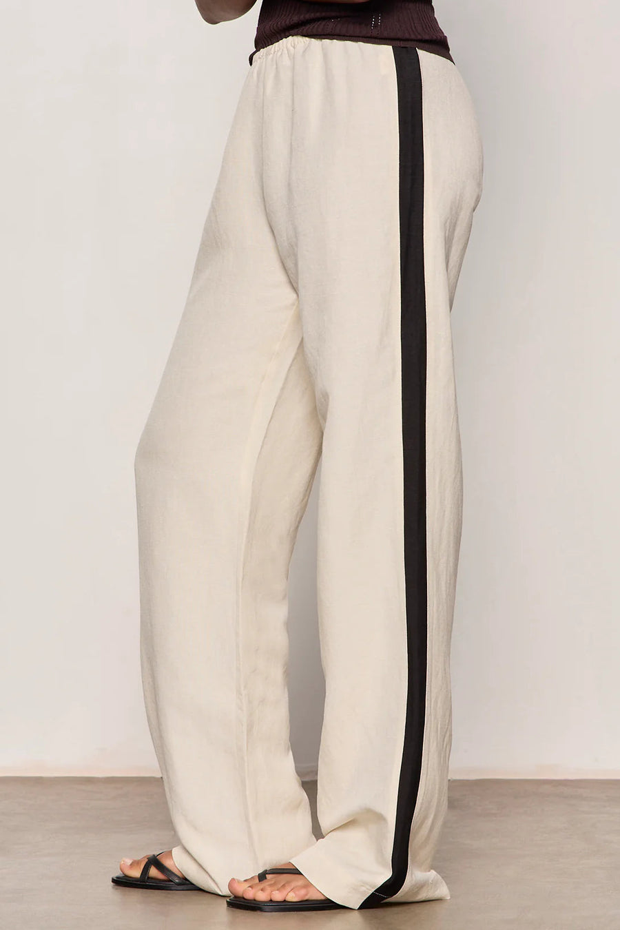 Woman wearing lightweight sand-colored wide-leg pants with contrasting black side stripe detailing. Features a comfortable pull-on elastic waistband, relaxed fit through the leg, and a fluid full-length silhouette. Designed with side pockets and a soft drape for effortless everyday wear, side