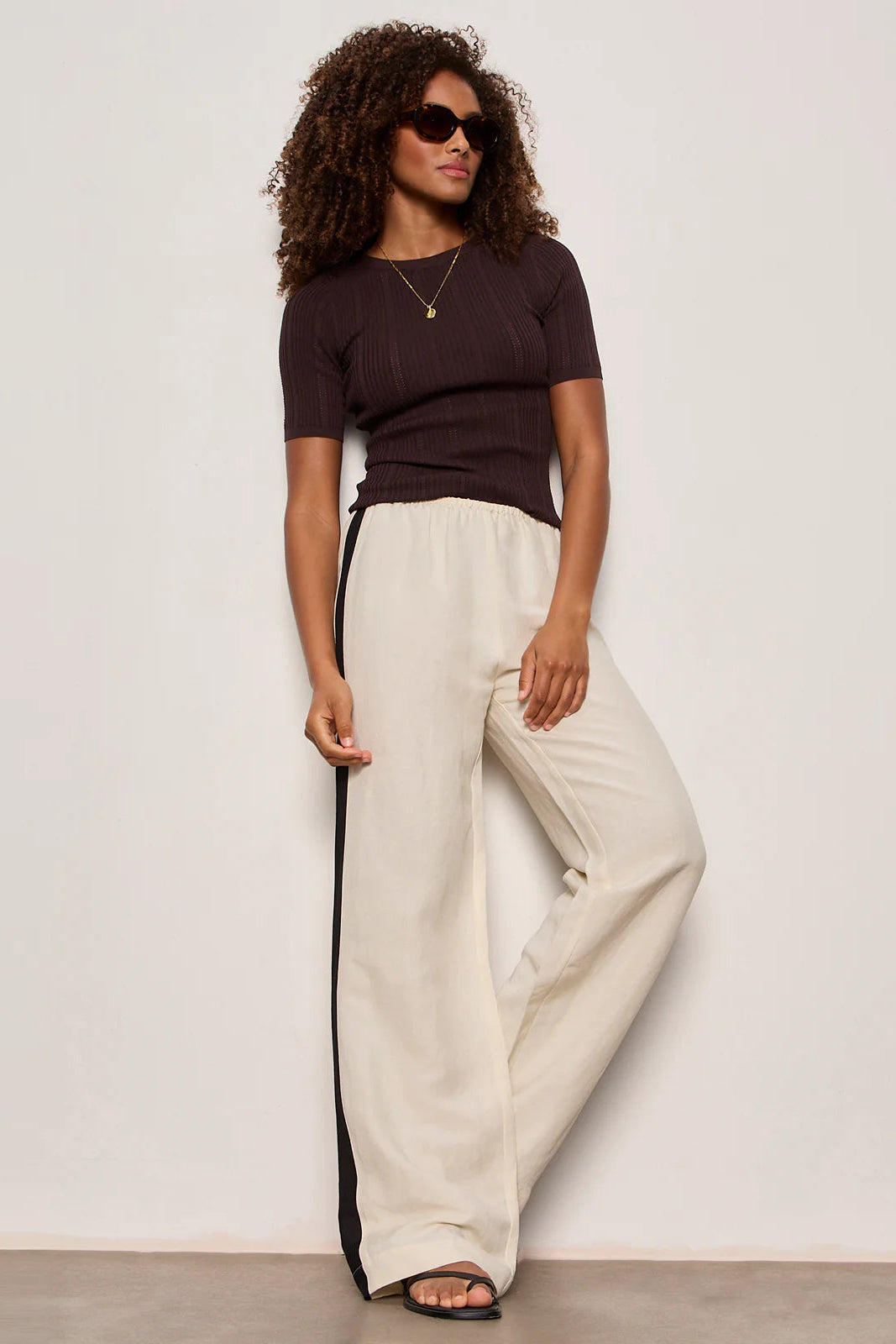 Woman wearing lightweight sand-colored wide-leg pants with contrasting black side stripe detailing. Features a comfortable pull-on elastic waistband, relaxed fit through the leg, and a fluid full-length silhouette. Designed with side pockets and a soft drape for effortless everyday wear, front