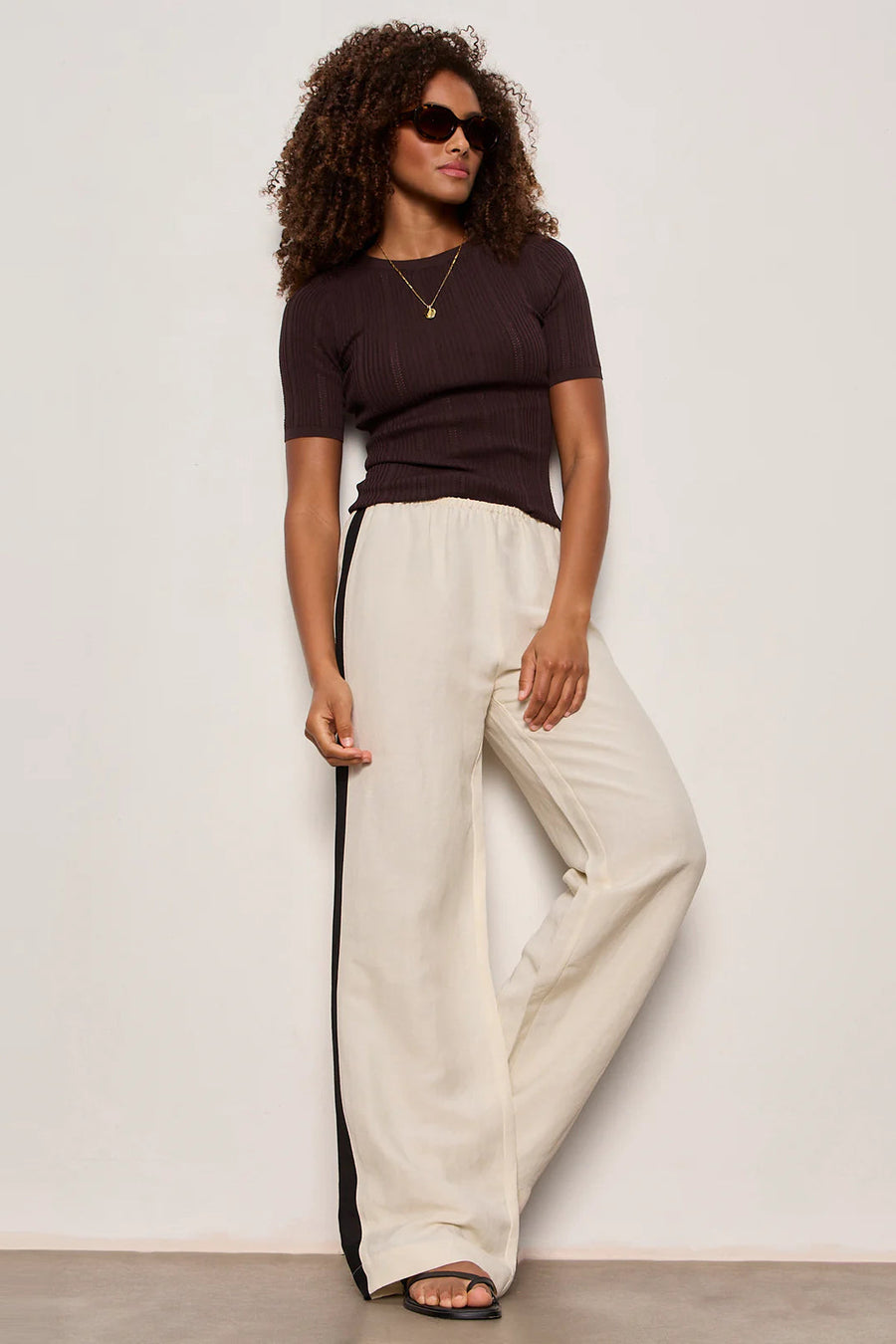 Woman wearing lightweight sand-colored wide-leg pants with contrasting black side stripe detailing. Features a comfortable pull-on elastic waistband, relaxed fit through the leg, and a fluid full-length silhouette. Designed with side pockets and a soft drape for effortless everyday wear, front