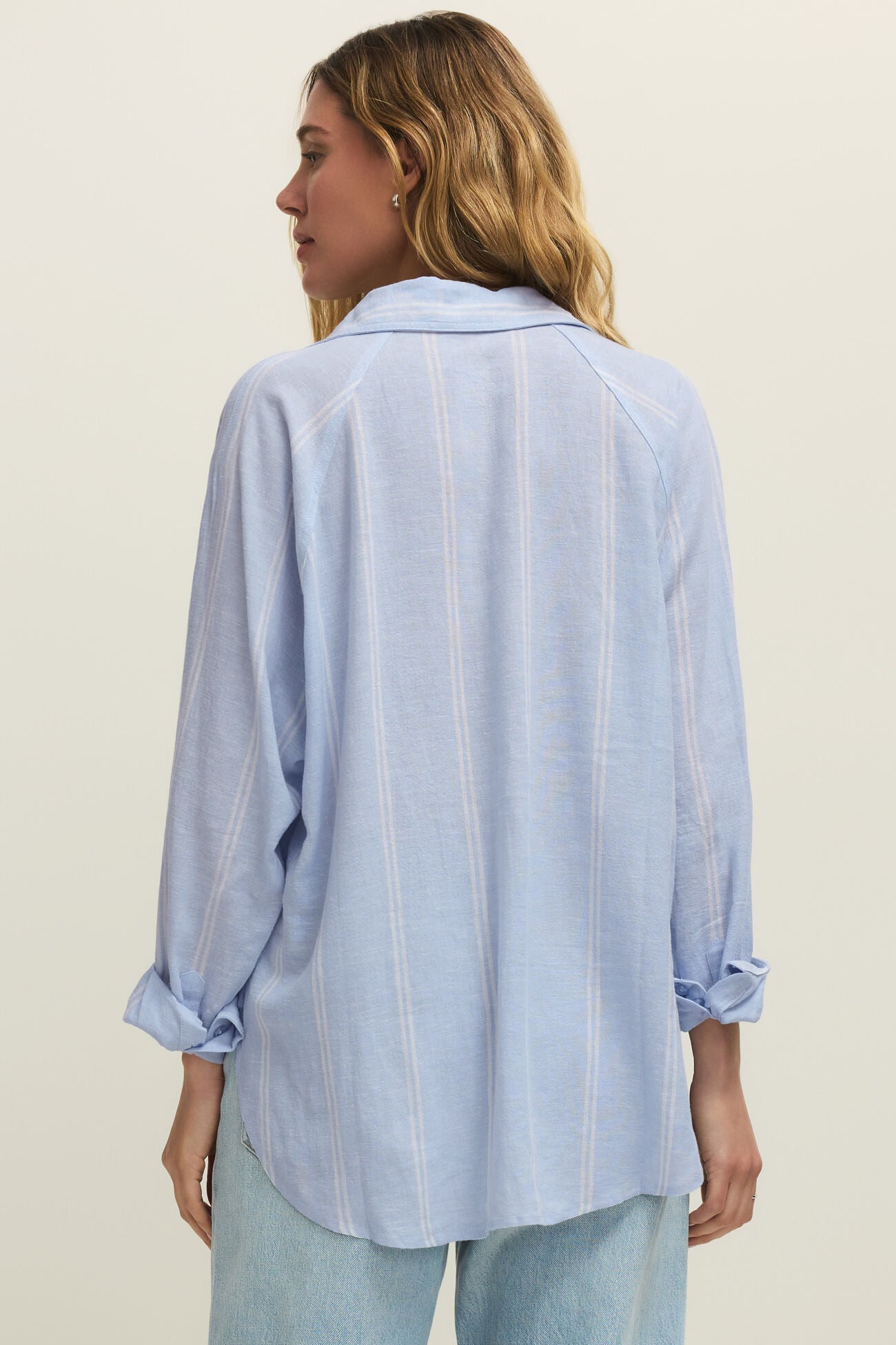 Woman wearing a oversized light blue long sleeve linen button down shirt with vertical white stripes, back view