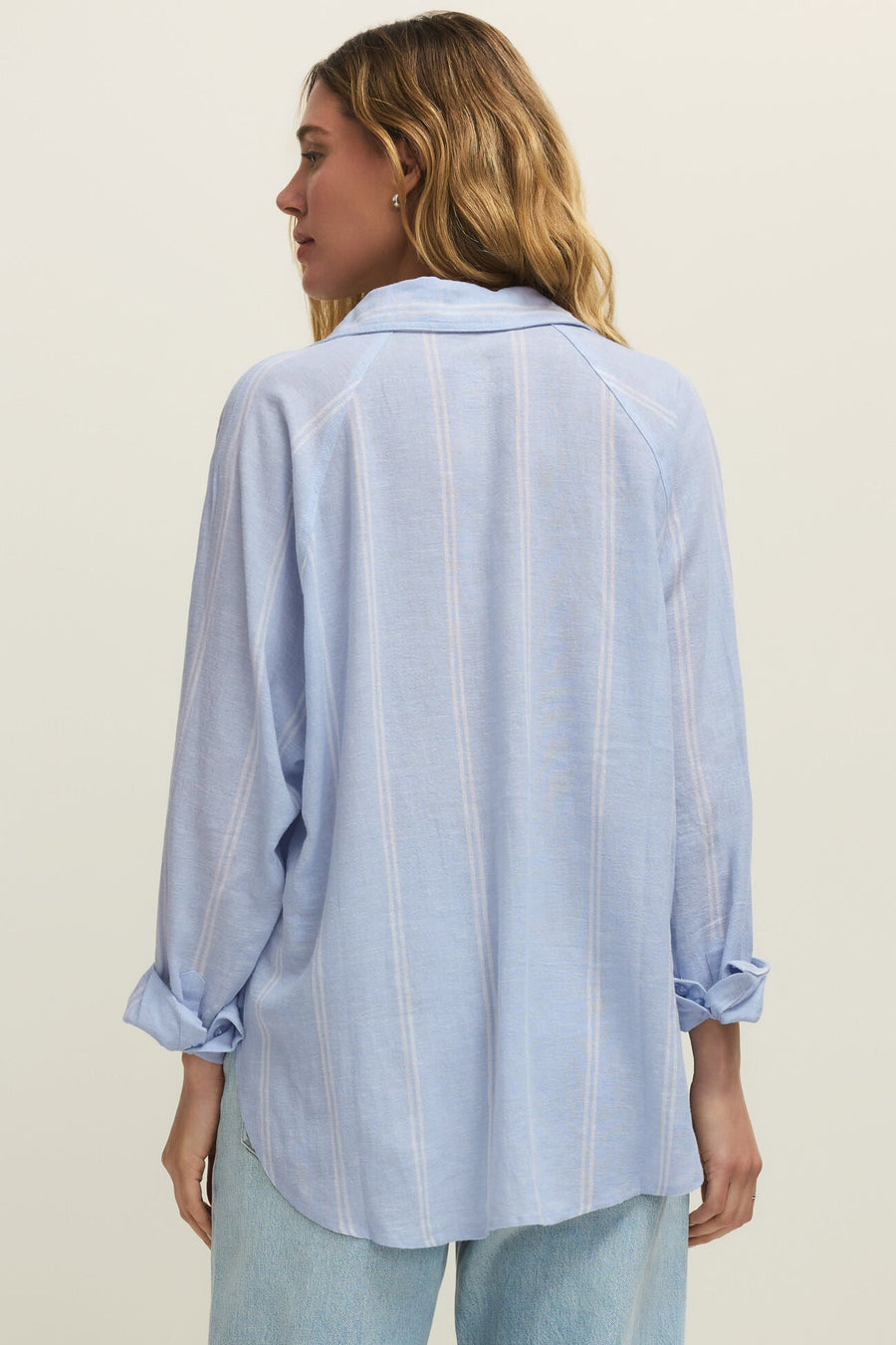 Woman wearing a oversized light blue long sleeve linen button down shirt with vertical white stripes, back view