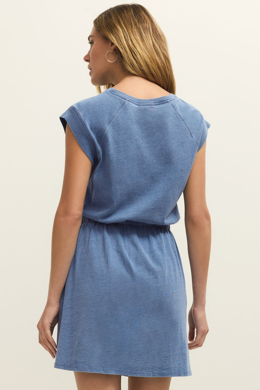 Woman wearing a sleeveless mid thigh jersey dress in a medium denim wash with drawstring at the waist, back view