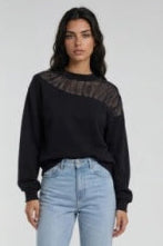 A model stands against a light gray studio background wearing a black long-sleeve top with sheer mesh shoulder panels,