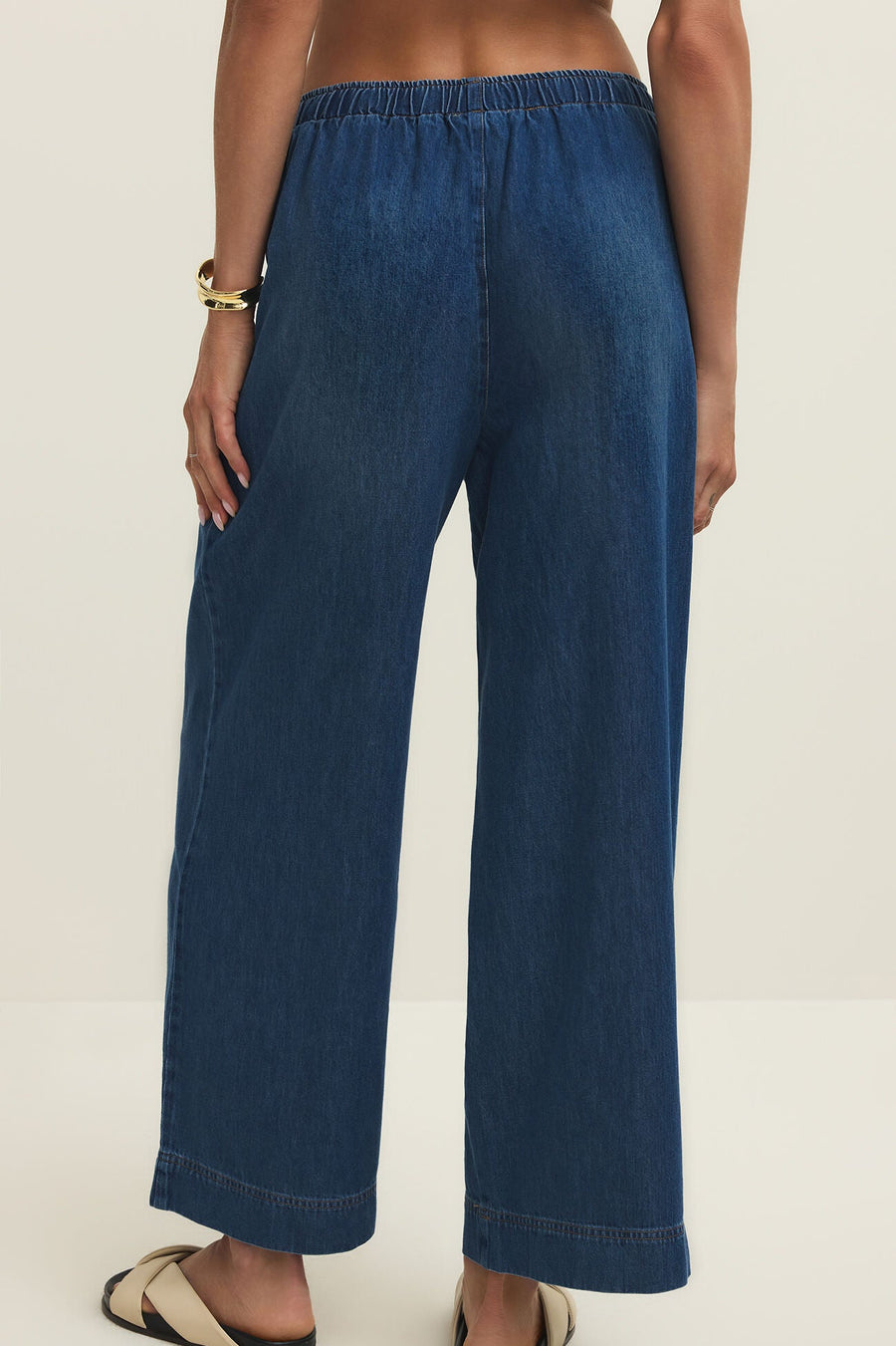 Woman wearing relaxed ankle length straight wide leg pants in a dark blue denim wash with a drawstring and pockets, back view